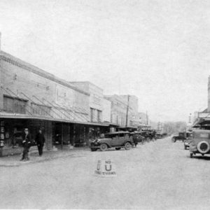 Old faded photo of men outside storefronts and parked cars with sign reading "Singer Drug Company Rexall Store" near hardware store "No U turn" road sign