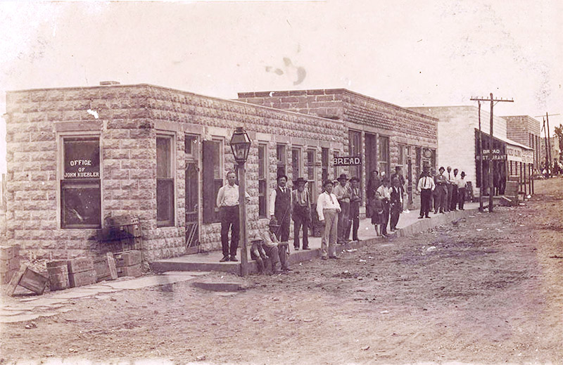 Group of white men standing on sidewalk outside single-story buildings on dirt street