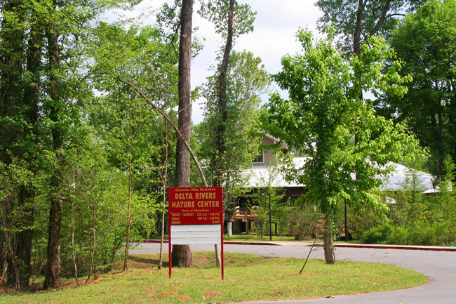 sign amid trees "Delta Rivers Nature Center" by road running in front of two-story structure