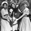Four white women in dresses hats and "Kennedy Johnson" belts pose feeding and petting donkey
