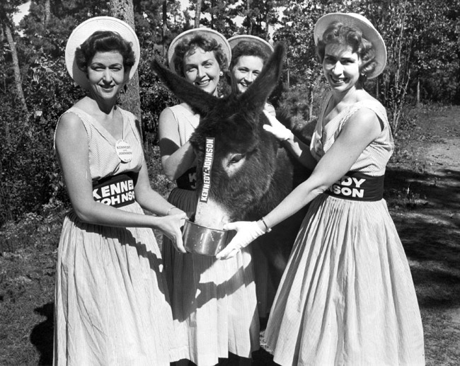 Four white women in dresses hats and "Kennedy Johnson" belts pose feeding and petting donkey