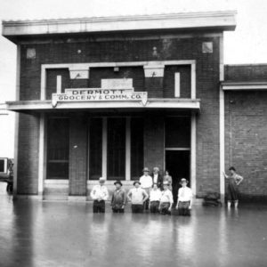 Nine people standing in a flooded street in front of a building labeled "Dermott Grocery & Comm. Co."
