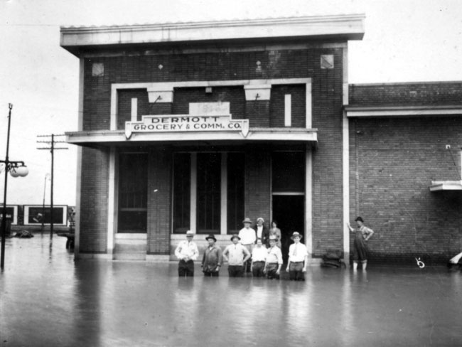Nine people standing in a flooded street in front of a building labeled "Dermott Grocery & Comm. Co."
