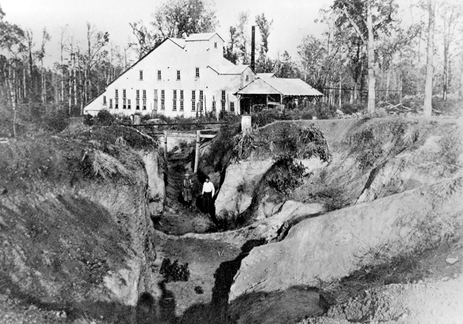 Exterior mine with two men standing, triangular building with smoke stack in background, forest
