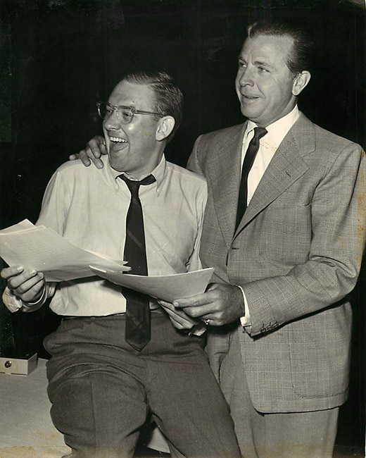 White man with glasses laughing next to white man in suit and tie