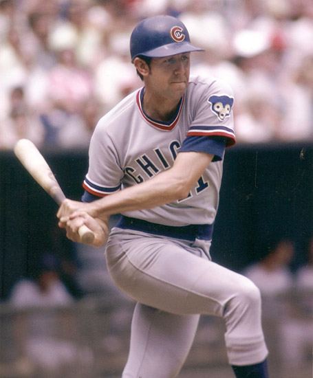 White man in baseball uniform with hard cap swinging a bat during a game with crowded stands in the background