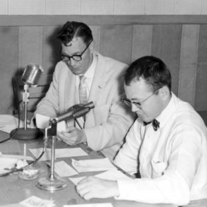 Two white men in formal attire at a desk with microphones reading from various handwritten letters.
