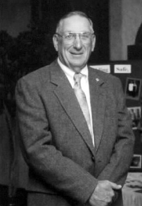 Portrait of older white man smiling with his hands crossed in suit tie glasses by table photo display