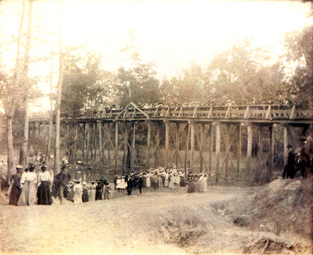 Group of people gathered at a river while spectators watch from a nearby wooden bridge