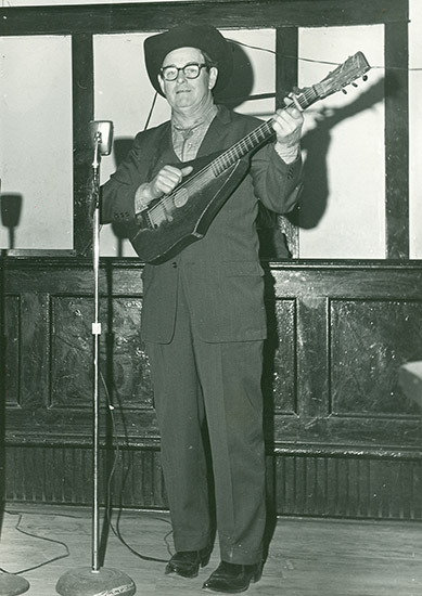 White man in suit and cowboy hat and glasses standing in front of a microphone stand and playing custom acoustic guitar
