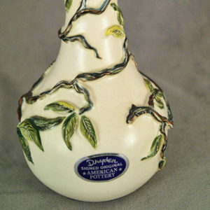 White vase with green leaves and vines wrapping around it and blue oval sticker on its front section
