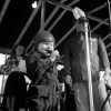 small child in leather helmet blows duck call at a microphone with adults standing in background
