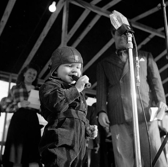 small child in leather helmet blows duck call at a microphone with adults standing in background