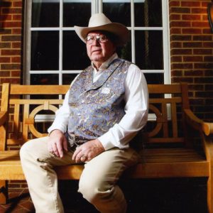 Old white man with glasses in western clothing and cowboy hat sitting on wooden bench outside brick building