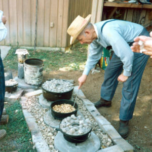 Old white man in overalls and hat cooking with three Dutch ovens in a fire pit on the ground