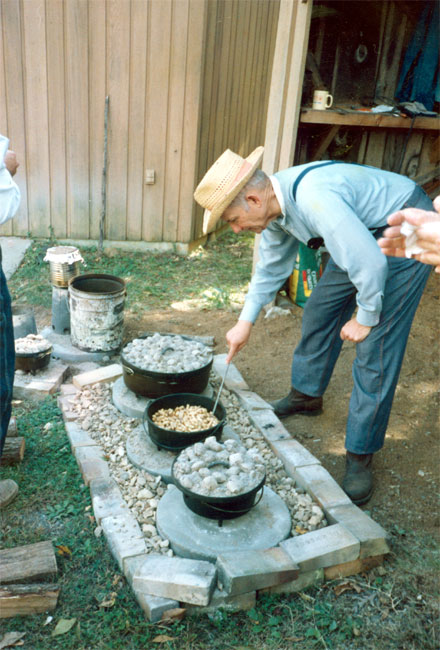 Old white man in overalls and hat cooking with three Dutch ovens in a fire pit on the ground