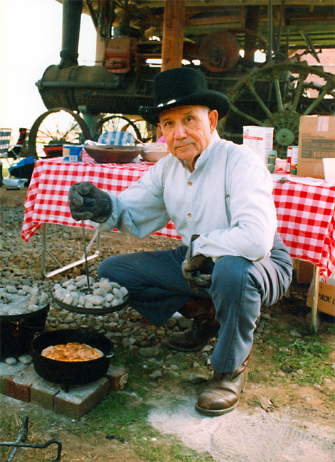 White man with hat posing while cooking with Dutch oven with a tractor and table behind him