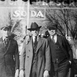 Three white men suits hats one with hand on friend's shoulder by "Soda" shop window