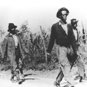White soldier with gun escorting group of young African-American men down dirt road