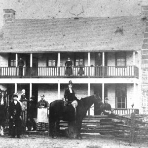 Various white people and two horses posing by large two-story building with chimneys on each end and wooden fence