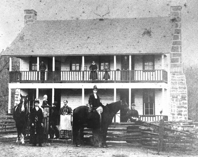 Various white people and two horses posing by large two-story building with chimneys on each end and wooden fence