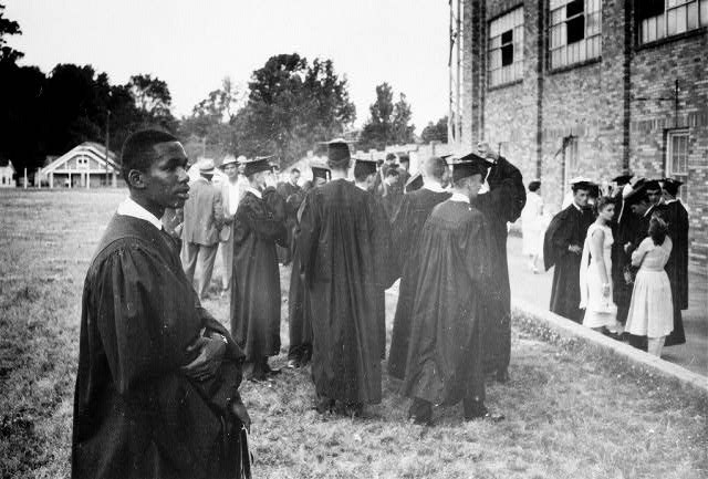 Black man in graduation gown outside brick stadium among graduates and people in suits dresses