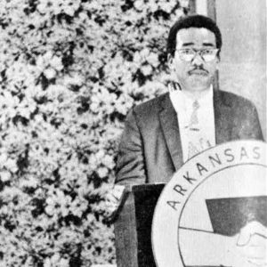 Young black man in suit tie glasses mustache at podium with round sign "Arkansas AFL"