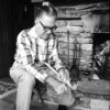 White man seated by stone hearth carving stick