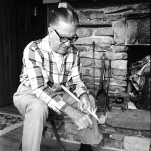White man seated by stone hearth carving stick