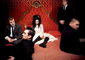 Young white woman in white dress with black hair and black knee-high boots lounging on red velvet bed with four white men in suits on or near the bed with red wallpaper behind them