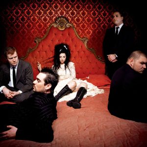 Young white woman in white dress with black hair and black knee-high boots lounging on red velvet bed with four white men in suits on or near the bed with red wallpaper behind them