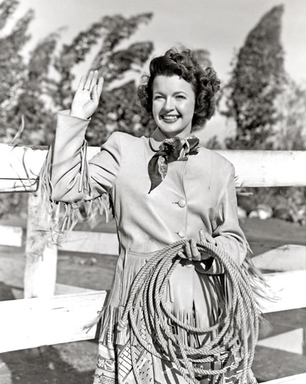White woman in western clothes holding a rope in left hand and raising her right in greeting