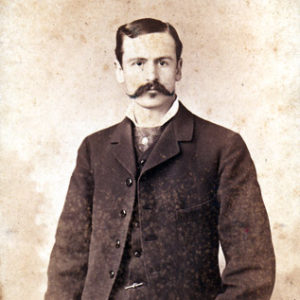 White man with handlebar mustache standing in three-piece suit and bow tie