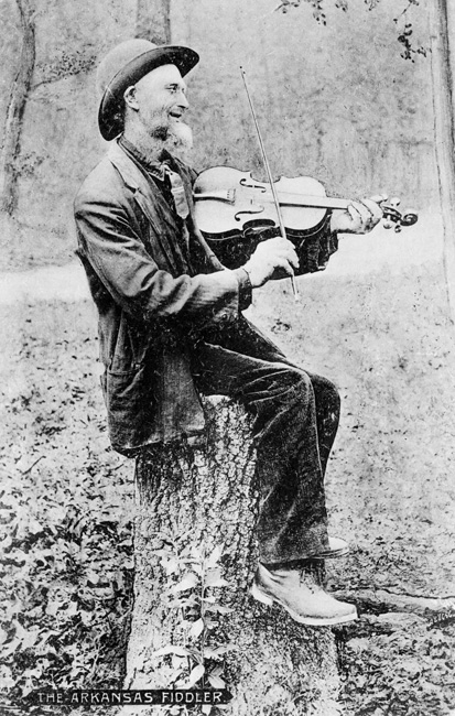 white man in suit and hat seated on tree stump playing fiddle, photo labeled "The Arkansas Fiddler"