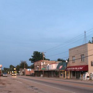 Parking lot and "Kyle's Barber Shop" building with sign across two-lane road from single and multistory brick storefronts