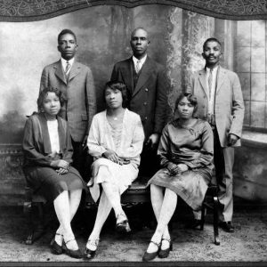 posed group photo of three black men in suits standing behind three seated black women in skirts on bench with legs crossed