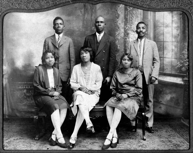 posed group photo of three black men in suits standing behind three seated black women in skirts on bench with legs crossed