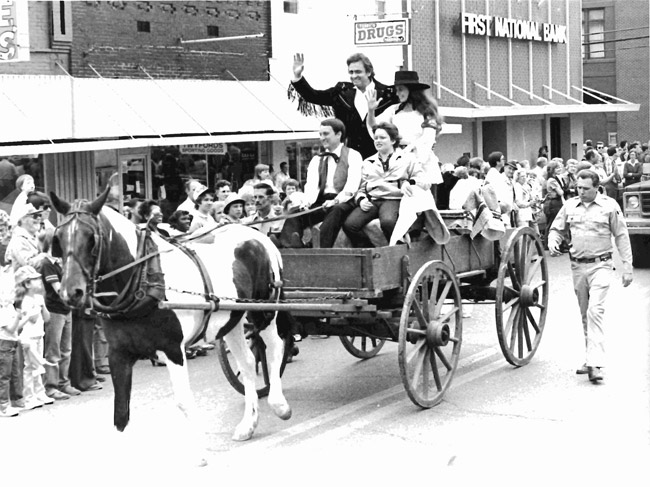 four white people in horse-drawn cart riding in parade down street lined with onlookers