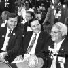 Three white men in suits smiling seated at political conference with people in background