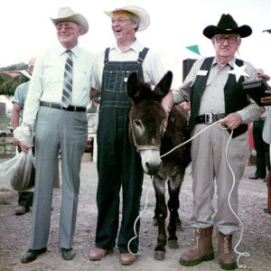 Three white men in cowboy hats pose with donkey and black woman judge at fair