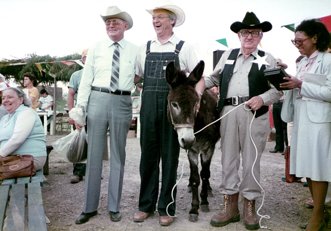 Three white men in cowboy hats pose with donkey and black woman judge at fair