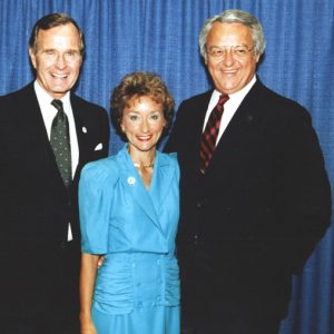 White woman in dress between two white men in suits pose smiling by curtain