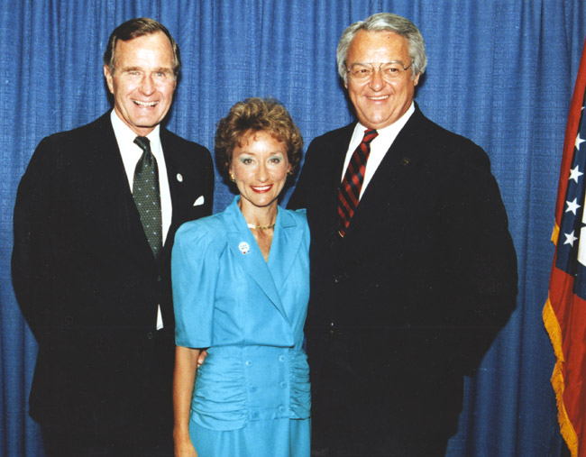 White woman in dress between two white men in suits pose smiling by curtain