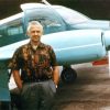 White man in pattern shirt and slacks posing by small airplane