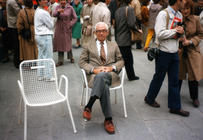White man posing in suit seated among standing white crowd