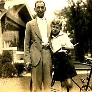 White man in suit hugging his son standing next to bicycle with house in the background