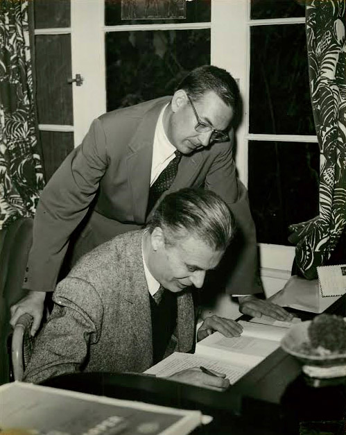 White man with glasses in suit standing over white man in suit working at his desk