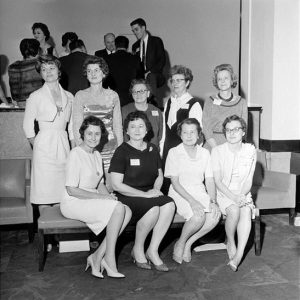 nine white women pose together with other people in background