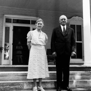 White woman in white dress and man in dark suit and tie pose on front steps with young girl watching through background doorway