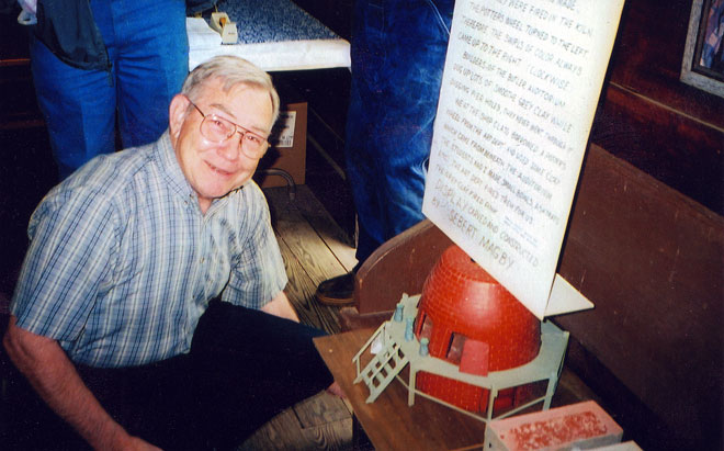 White man kneeling by small conical brick home model and text display on bench